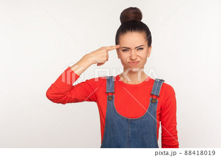 Crazy cuckoo mind. Portrait of girl with hair bun in denim overalls showing stupid gesture and looking with displeased annoyed face, accusing insane idea. studio shot isolated on white background Crazy cuckoo mind. Portrait of girl with hair bun in denim overalls showing stupid gesture and looking with displeased annoyed face, accusing insane idea. studio shot isolated on white background 88418019