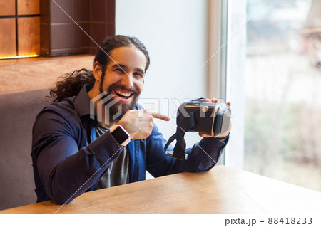 Portrait of happy bearded young adult man in casual style sitting in cafe, holding vr and pointing finger on simulator with happy face, looking at camera and toothy smile. Indoor, lifestyle concept Portrait of happy bearded young adult man in casual style sitting in cafe, holding vr and pointing finger on simulator with happy face, looking at camera and toothy smile. Indoor, lifestyle concept 88418233