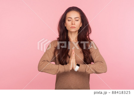 Meditation, peaceful mind. Portrait of calm concentrated young woman with brunette wavy hair holding hands in namaste gesture and relaxing, doing yoga. indoor studio shot isolated on pink background 88418325