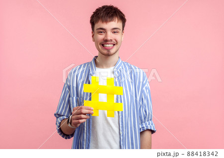 Popularity in social media. Portrait of happy young brown-haired man in casual shirt holding large big yellow hashtag sign, grinning with excitement. indoor studio shot isolated on pink background Popularity in social media. Portrait of happy young brown-haired man in casual shirt holding large big yellow hashtag sign, grinning with excitement. indoor studio shot isolated on pink background 88418342