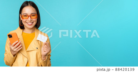 Online shopping. Stylish young asian woman in sunglasses, showing credit card and using smartphone, paying in internet, making purchase, standing over blue background Online shopping. Stylish young asian woman in sunglasses, showing credit card and using smartphone, paying in internet, making purchase, standing over blue background 88420139