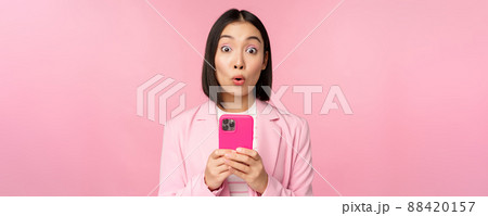 Portrait of asian businesswoman with surprised face, using smartphone app, wearing business suit. Korean girl with mobile phone and excited face expression, pink background 88420157