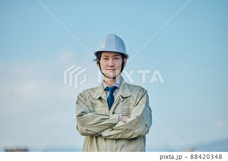 青空を背景に腕組みをして立つ作業服姿の若い男性 88420348