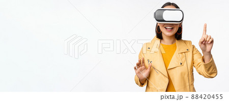 Image of young asian woman wearng VR headset, using virtual reality glasses and smiling, pointing at smth, choosing smth, standing over white background Image of young asian woman wearng VR headset, using virtual reality glasses and smiling, pointing at smth, choosing smth, standing over white background 88420455