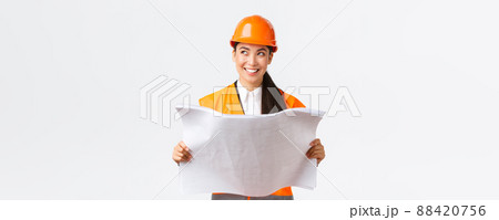 Creative asian female architect imaging her plan, standing in safety helmet and looking away thoughtful while reading blueprints, picturing buiding after finish construction works, white background Creative asian female architect imaging her plan, standing in safety helmet and looking away thoughtful while reading blueprints, picturing buiding after finish construction works, white background 88420756