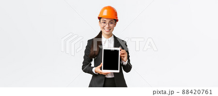 Smiling professional female engineer, saleswoman or estate agent showing location to client, standing in safety helmet and business suit, introduce construction project at tablet screen Smiling professional female engineer, saleswoman or estate agent showing location to client, standing in safety helmet and business suit, introduce construction project at tablet screen 88420761