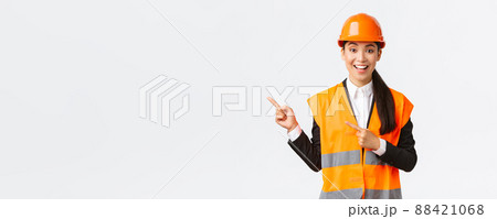 Building, construction and industrial concept. Smiling female engineer in helmet and reflective clothing showing way, pointing upper left corner, talking about constructing project, white background Building, construction and industrial concept. Smiling female engineer in helmet and reflective clothing showing way, pointing upper left corner, talking about constructing project, white background 88421068