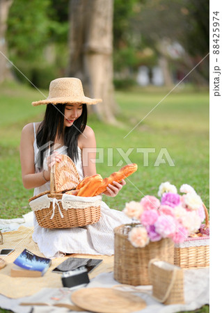 Woman with a pretty dress, wicker picnic basket with baguette and accessories on picnic cloth. 88425974