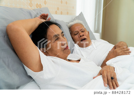 Older Asian woman laying open her eyes in bed beside her husband who snores and Makes noise, Marital problems. Older Asian woman laying open her eyes in bed beside her husband who snores and Makes noise, Marital problems. 88428172