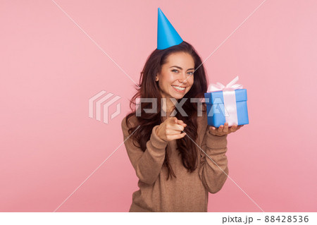 This present for you. Portrait of happy joyful young woman wearing party cone on head holding box and pointing to camera, holiday tradition of gift giving. studio shot isolated on pink background 88428536