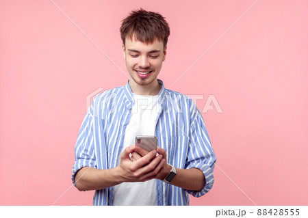 Portrait of positive brown-haired man with small beard and mustache in casual striped shirt holding cellphone, smiling while reading message, good news. indoor studio shot isolated on pink background Portrait of positive brown-haired man with small beard and mustache in casual striped shirt holding cellphone, smiling while reading message, good news. indoor studio shot isolated on pink background 88428555