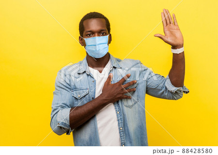 I swear. Portrait of honest serious man in denim casual shirt with surgical medical mask keeping hand on chest, raising palm, giving promise, pledge. indoor studio shot isolated on yellow background 88428580