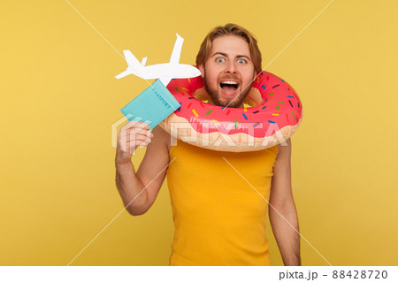 Delighted tourist guy in undershirt standing with pink donut rubber ring, holding passport document and airplane mockup, looking at camera, shouting rejoicing travel tour. indoor studio shot isolated 88428720