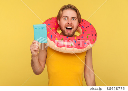 Happy optimistic funny tourist guy in undershirt standing with pink donut rubber ring around head and holding passport document, visa free travel abroad. studio shot isolated on yellow background Happy optimistic funny tourist guy in undershirt standing with pink donut rubber ring around head and holding passport document, visa free travel abroad. studio shot isolated on yellow background 88428776