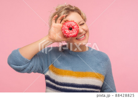 Sweet dessert, confection. Portrait of cheerful woman with short curly hair in sweater holding doughnut, looking through donut at camera and smiling, having fun. indoor studio shot pink background 88428825