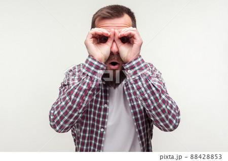 Portrait of curious wondered bearded man in casual plaid shirt looking through fingers gesturing binoculars, amazed surprised by what he discovers. indoor studio shot isolated on white background 88428853