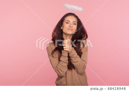 Portrait of lovely sexy young woman with halo above head looking at camera with alluring smile and holding hands in prayer, asking and seducing with angelic beauty. indoor studio shot pink background 88428948