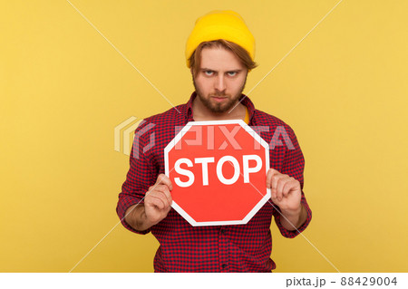 Determined serious hipster guy in beanie hat and checkered shirt holding traffic Stop sign and looking menace, warning of road safety, pedestrian crossing. studio shot isolated on yellow background Determined serious hipster guy in beanie hat and checkered shirt holding traffic Stop sign and looking menace, warning of road safety, pedestrian crossing. studio shot isolated on yellow background 88429004