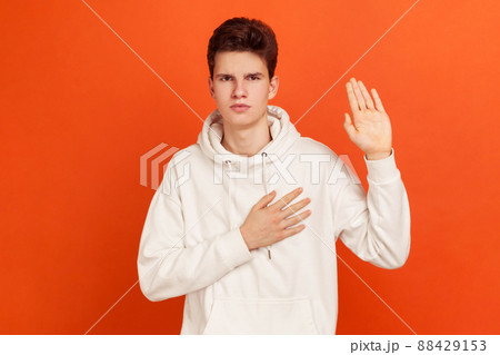 Serious patriotic male in white sweatshirt with hood holding hand on heart, juryman swearing to speak truth in court, honor and conscience. Indoor studio shot isolated on orange background 88429153