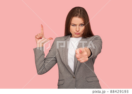 You are fired. Portrait of displeased young woman in business suit pointing to camera and making L sign with fingers, showing loser gesture, accusing for failure. studio shot, pink background 88429193