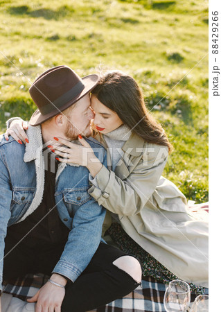 Man and woman in fashion clothes sitting on a nature on a picnic rug. Man wearing jacket and a black hat and woman coat. Woman hugging a man. 88429266
