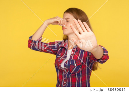 Bad smell. Portrait of displeased ginger girl in checkered shirt pinching nose and showing stop gesture, grimacing in disgust, avoiding stinky odor. indoor studio shot isolated on yellow background 88430015