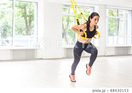 Portrait of young fit woman in black sportswear training arms with trx fitness straps in the gym doing push ups train upper body chest shoulders pecs triceps with raised leg. Indoor, window background 88430153