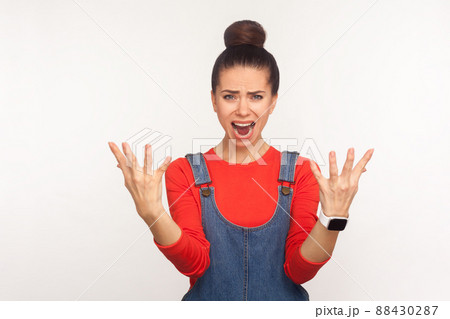 How could you. Portrait of irritated girl with hair bun in denim overalls screaming with anger, raising hands in question why, what reason, misunderstanding in conflict. studio shot white background 88430287
