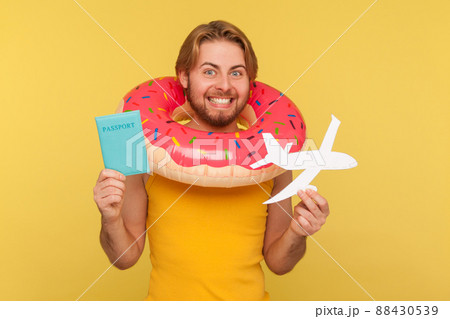 Excited optimistic funny tourist guy in undershirt standing with pink donut rubber ring, holding passport document and airplane mockup, rejoicing travel tour. studio shot isolated on yellow background 88430539
