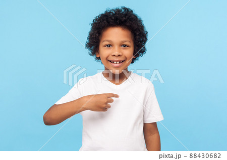 This is me. Portrait of happy preschool curly boy in T-shirt joyfully looking at camera and pointing to himself, proud of own success, egoistic child. indoor studio shot isolated on blue background 88430682