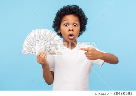 Wow, I'm rich. Amazed lucky wealthy little boy with curly hair pointing to fan of dollar banknotes, shocked by lots of cash and looking at camera with astonishment. indoor studio shot, blue background Wow, I'm rich. Amazed lucky wealthy little boy with curly hair pointing to fan of dollar banknotes, shocked by lots of cash and looking at camera with astonishment. indoor studio shot, blue background 88430692