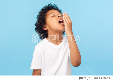 Portrait of sleepy tired little boy with curly hair in white T-shirt covering mouth with hand while yawning with eyes closed, drowsy child waking up. indoor studio shot isolated on blue background Portrait of sleepy tired little boy with curly hair in white T-shirt covering mouth with hand while yawning with eyes closed, drowsy child waking up. indoor studio shot isolated on blue background 88431024