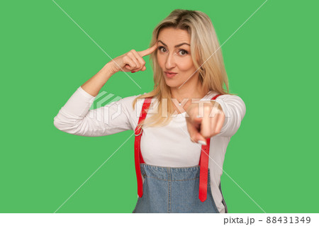 You're idiot with insane crazy plan. Portrait of displeased adult woman in denim overalls showing stupid gesture and pointing to camera, accusing of dumb idea. studio shot isolated on green background You're idiot with insane crazy plan. Portrait of displeased adult woman in denim overalls showing stupid gesture and pointing to camera, accusing of dumb idea. studio shot isolated on green background 88431349