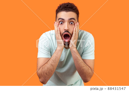 Unbelievable shocking news. Portrait of stunned brunette man with beard in casual white t-shirt holding face and keeping mouth wide open in amazement. indoor studio shot isolated on orange background Unbelievable shocking news. Portrait of stunned brunette man with beard in casual white t-shirt holding face and keeping mouth wide open in amazement. indoor studio shot isolated on orange background 88431547