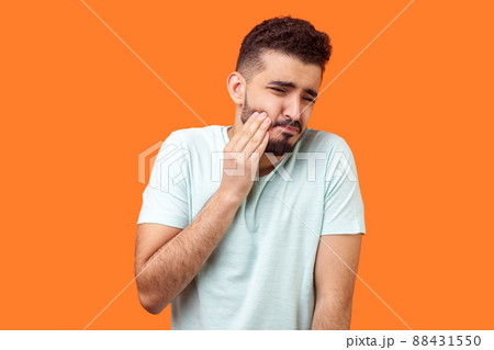 Dental problem. Portrait of unhealthy brunette man with beard in white t-shirt touching his cheek and grimacing from terrible toothache, sensitive teeth. indoor studio shot isolated, orange background Dental problem. Portrait of unhealthy brunette man with beard in white t-shirt touching his cheek and grimacing from terrible toothache, sensitive teeth. indoor studio shot isolated, orange background 88431550