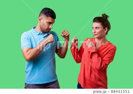 Come on let's fight. Portrait of lovely couple in casual wear standing together, keeping fists clenched and looking at each other ready to fight, joke. isolated on green background, indoor studio shot Come on let's fight. Portrait of lovely couple in casual wear standing together, keeping fists clenched and looking at each other ready to fight, joke. isolated on green background, indoor studio shot 88431551