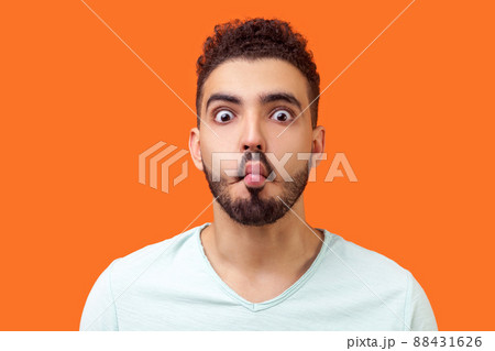 Portrait of funny silly brunette man with beard in casual white t-shirt looking at camera and making fish face with lips and big eyes, fooling around. indoor studio shot isolated on orange background Portrait of funny silly brunette man with beard in casual white t-shirt looking at camera and making fish face with lips and big eyes, fooling around. indoor studio shot isolated on orange background 88431626