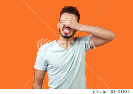 Don't want to look at this. Portrait of depressed brunette man with beard in white t-shirt covering eyes, ignoring problems with angry scared face. indoor studio shot isolated on orange background Don't want to look at this. Portrait of depressed brunette man with beard in white t-shirt covering eyes, ignoring problems with angry scared face. indoor studio shot isolated on orange background 88431659