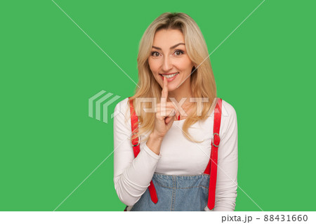 Shh, don't speak. Portrait of positive adult blond woman in stylish overalls holding finger on lips in silence gesture, asking to keep secret, shushing about conspiracy. indoor studio shot, isolated 88431660
