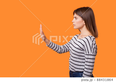 Side view portrait of worried young woman with brown hair in long sleeve striped shirt standing, raising arm showing stop gesture, warning sign. indoor studio shot isolated on orange background 88431864