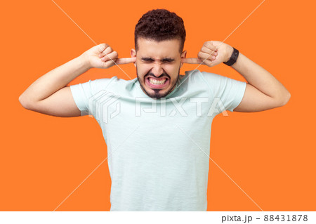 Don't want to listen anymore. Portrait of brunette man standing with irritated grimace, tightly covering his ears not to hear annoying loud sound. indoor studio shot isolated on orange background 88431878