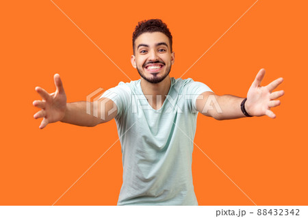 Come into my arms. Portrait of good natured, extremely happy brunette man with beard in white t-shirt reaching out to camera, stretching arms to hug you. studio shot isolated on orange background 88432342