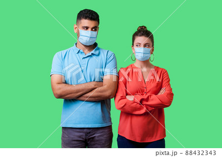 Portrait of upset couple with surgical medical mask standing together with crossed hands, looking sideways at each other with resentful glance, suspicion. isolated on green background, indoor studio 88432343