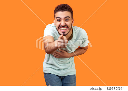 Portrait of extremely happy brunette man with beard in white t-shirt holding hand on belly, pointing at camera and laughing out loud, making fun of you. studio shot isolated on orange background 88432344