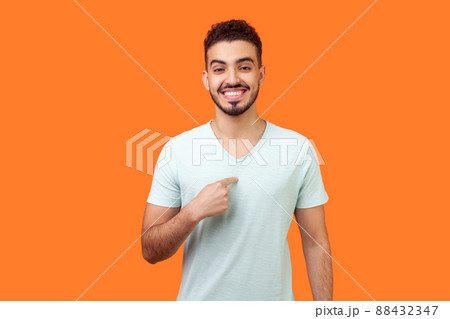 This is me. Portrait of glad satisfied brunette man with beard in white t-shirt pointing at himself, looking at camera with proud and boastful smile. indoor studio shot isolated on orange background 88432347