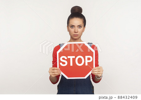 Attention, prohibited way. Portrait of serious girl with hair bun holding octagonal Stop symbol, showing red traffic sign, warning of restricted area. indoor studio shot isolated on white background 88432409