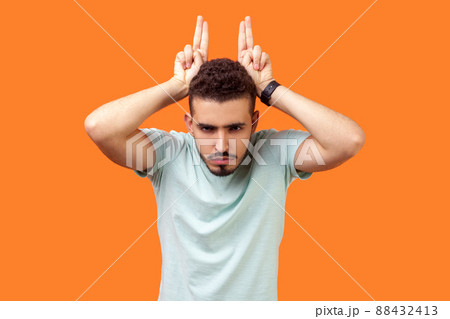 Portrait of angry bully brunette man with beard in white t-shirt showing bull horn gesture with fingers over head, looking hostile and threatening. indoor studio shot isolated on orange background 88432413