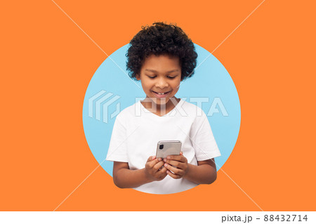 Happy excited boy with curly hair reading message on smartphone and smiling, satisfied with children mobile application, using cellphone. indoor shot isolated in a round hole on orange background 88432714