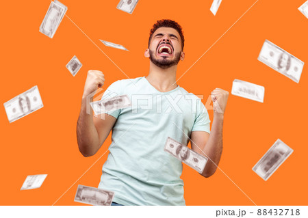 Money rain, winner and rich. Bottom view of ecstatic motivated man standing with raised fists and shouting for joy, winner excited for success. dollars falling. indoor isolated on orange background 88432718