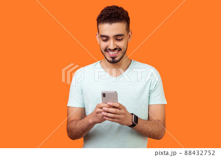 Portrait of cheerful brunette man with beard in white t-shirt using cellphone and smiling, reading good news message, enjoying mobile application. indoor studio shot isolated on orange background 88432752